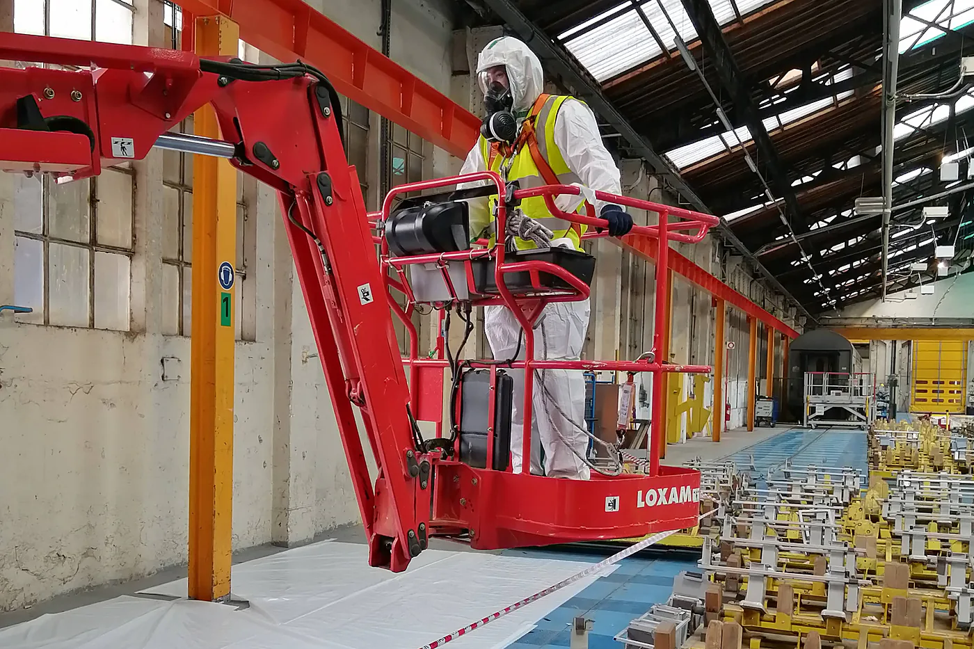 Opérateur en combinaison intégrale et masque complet sur une nacelle élévatrice, dans un hall industriel à la zone balisée