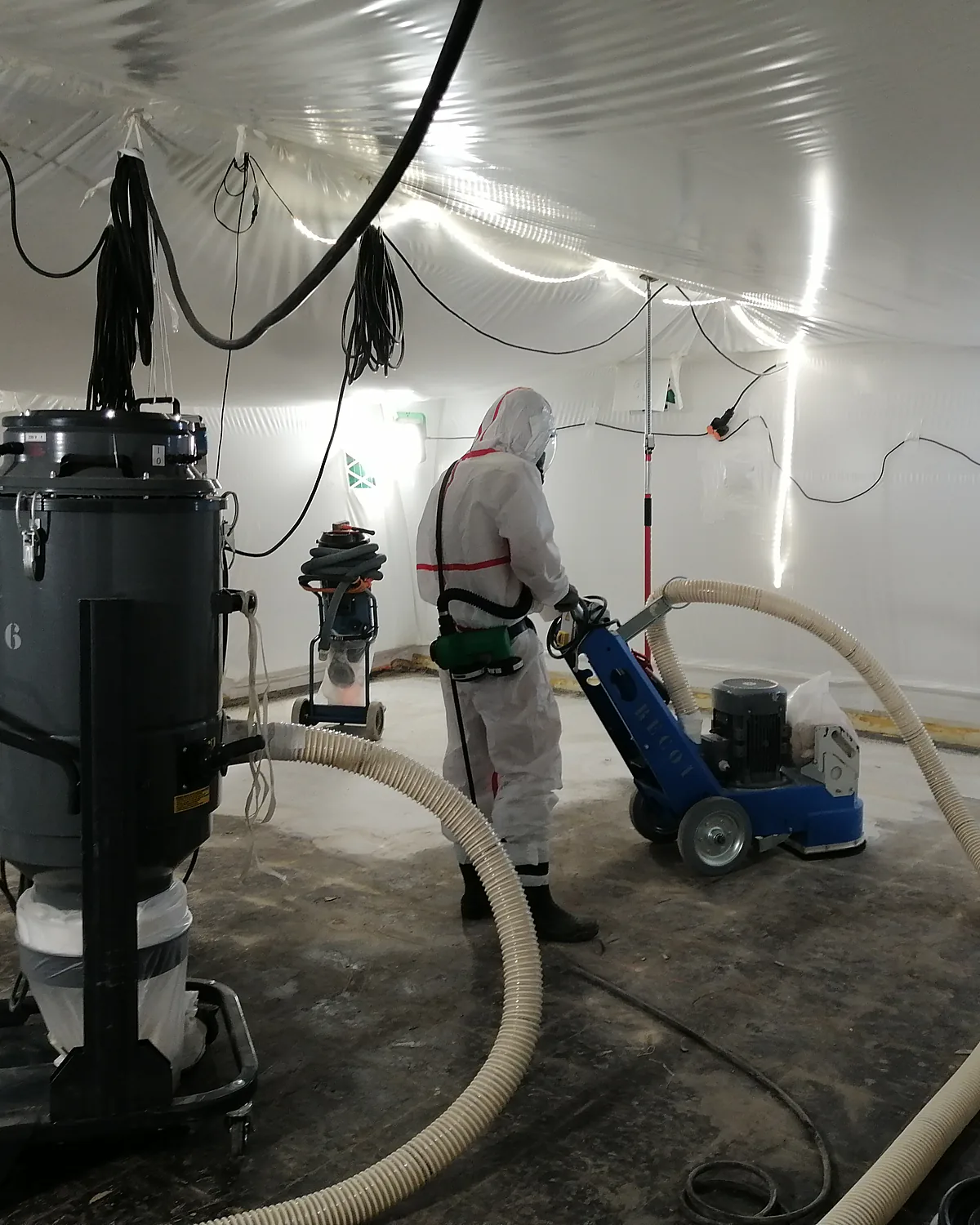 Opérateur en combinaison et masque respiratoire ponçant un sol dans une zone confinée, aspirateurs à filtre absolu en place