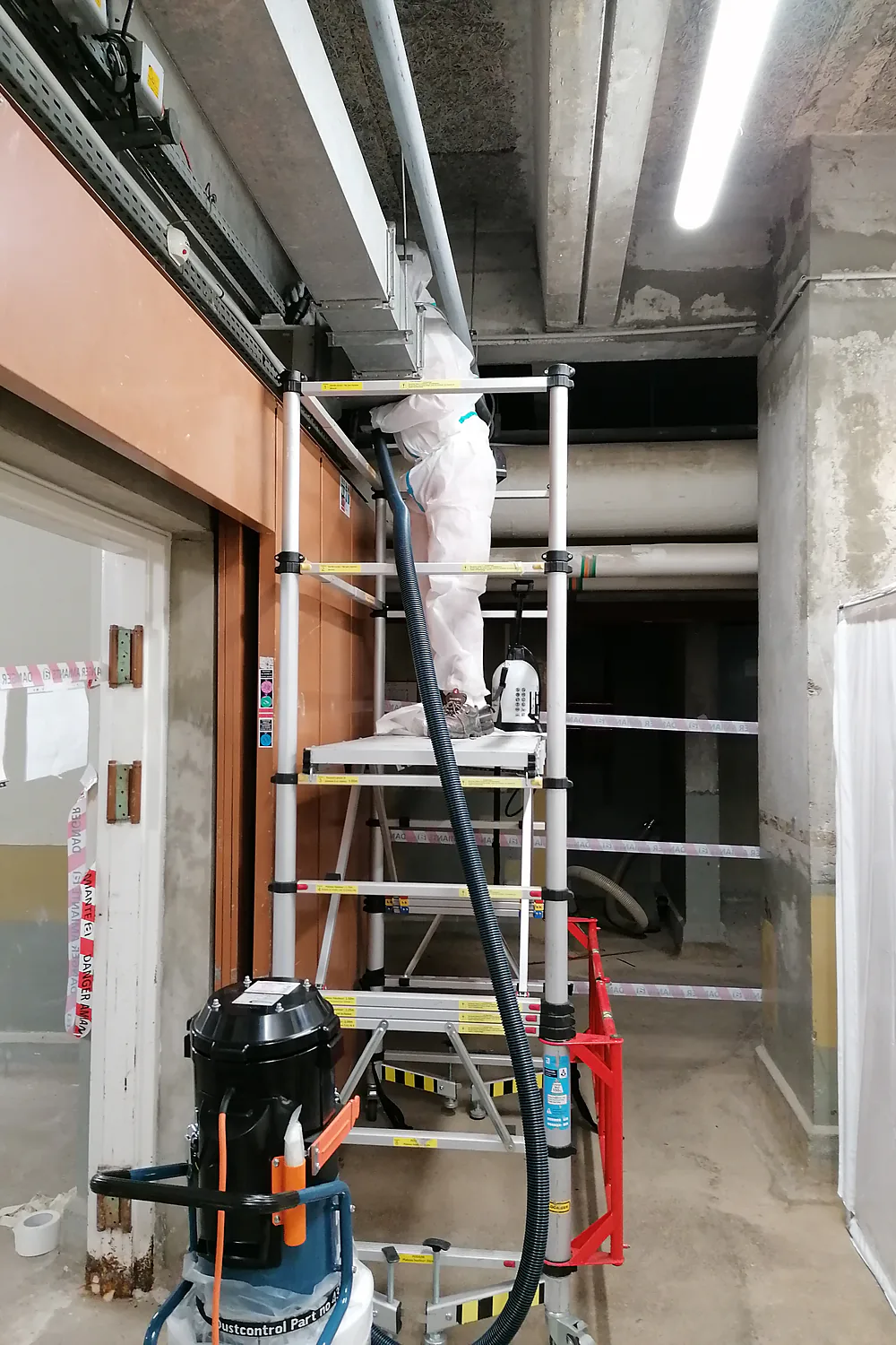 Opérateur en combinaison sur un échafaudage roulant, traitant le plafond d'un sous-sol avec un aspirateur à filtre absolu